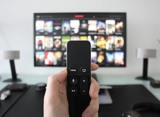 A person holds a remote control in front of a TV displaying a streaming platform interface with various movie and show thumbnails. The room includes a desk with two small lamps on either side of the TV.