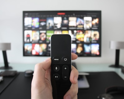 A person holds a remote control in front of a TV displaying a streaming platform interface with various movie and show thumbnails. The room includes a desk with two small lamps on either side of the TV.