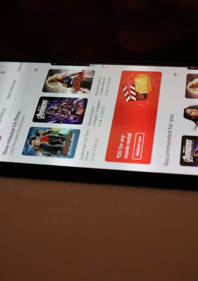 A smartphone displaying a streaming app interface, showing various movie and TV show thumbnails. The screen includes categories like 'For you' and 'New release hit films', with images from popular films. The background is a dimly lit setting with a fabric surface.