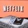 A screen displaying the Netflix logo in bold, black and white lettering against a vibrant red background. In front of the screen, there is a closed box for the board game 'Sequence', with a smartphone and wireless earbuds resting on top.