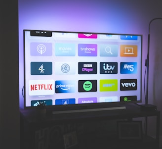 A flat-screen television is displayed in a dimly lit room, showcasing a variety of streaming service apps on its screen, such as Netflix, Prime Video, and Spotify. The TV is positioned on a black stand beside a tall floor lamp, casting a soft ambient light on the surrounding area.