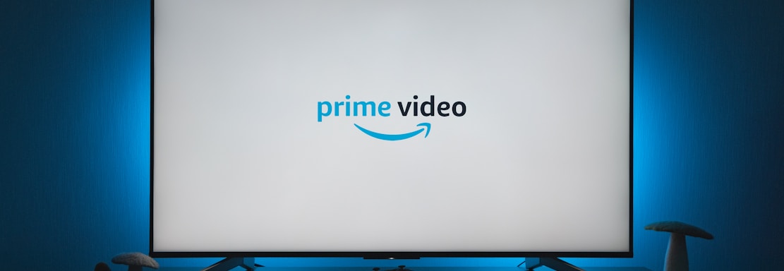 A television screen displays the Prime Video logo, situated on an entertainment unit in a dimly lit room. The room is illuminated with a subtle blue backlight, creating a calming atmosphere. Two small decorative items, resembling mushrooms, are placed on either side of the unit.