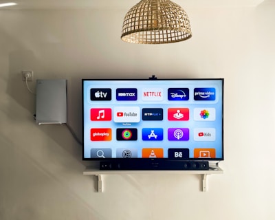 A modern living room features a wall-mounted flat-screen television displaying multiple streaming app icons, such as Apple TV, HBO Max, Netflix, Disney+, and Prime Video. Below the TV is a soundbar sitting on a small white shelf. A wicker pendant light hangs from the ceiling above, and the wall is painted a neutral color.