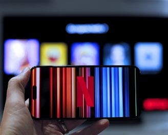 A hand holding a smartphone displaying the Netflix logo on its screen with a background of colorful vertical lines. In the background, a television screen shows blurry icons or profiles with various colors.