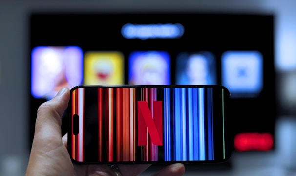 A hand holding a smartphone displaying the Netflix logo on its screen with a background of colorful vertical lines. In the background, a television screen shows blurry icons or profiles with various colors.