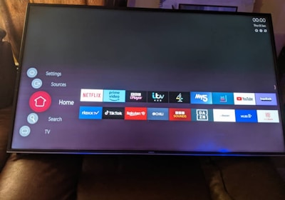 A television screen displays a smart TV interface with multiple app icons arranged in rows. Options such as Settings, Sources, Home, Search, and TV are listed on the left side. Some recognizable app icons include Netflix, Prime Video, BBC iPlayer, ITV Hub, All 4, My5, YouTube, and TikTok. The time and date are shown in the top right corner.