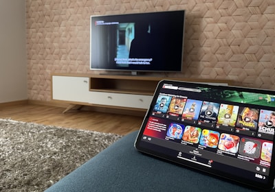 A modern living room features a flat-screen television mounted on a light-colored wooden stand with a patterned wall backdrop. Below the TV, a carpet lies on a wooden floor. In the foreground, a tablet displays a streaming platform interface with various show thumbnails, suggesting a relaxed entertainment setup.